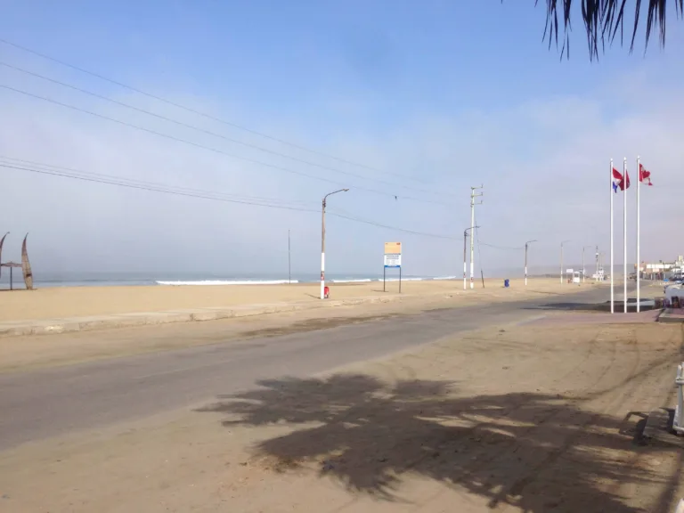 Sandiger Küstenweg mit Palmenschatten, Fahnenmasten und Strand im Hintergrund