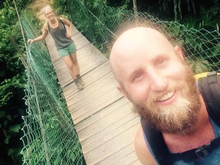Selfie eines bärtigen Mannes auf einer Hängebrücke, hinter ihm läuft eine Frau