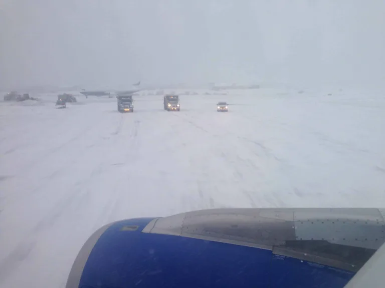 Blick aus dem Flugzeugfenster auf ein verschneites Flughafenvorfeld mit Fahrzeugen