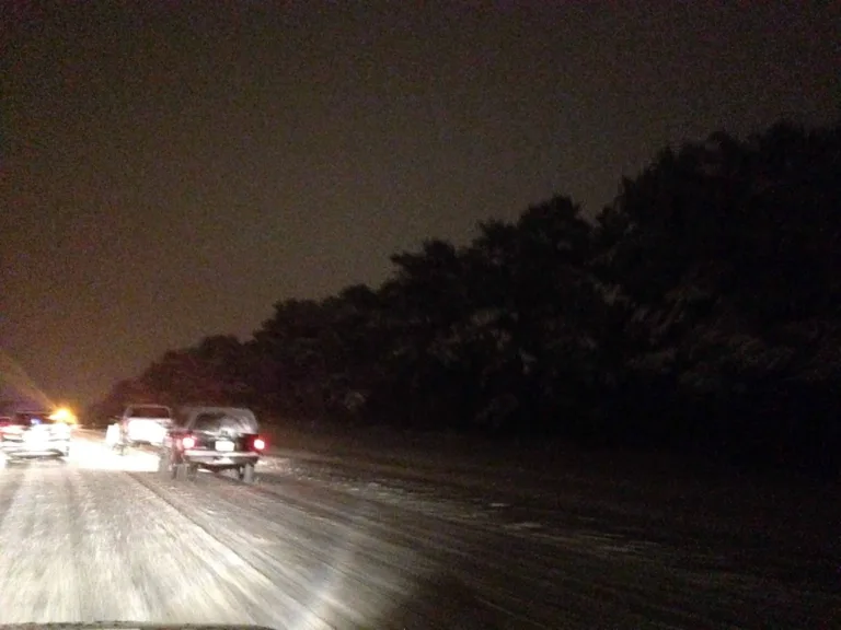 Verschneite Fahrbahn bei Nacht mit stehenden Autos und dunklem Wald am Strassenrand